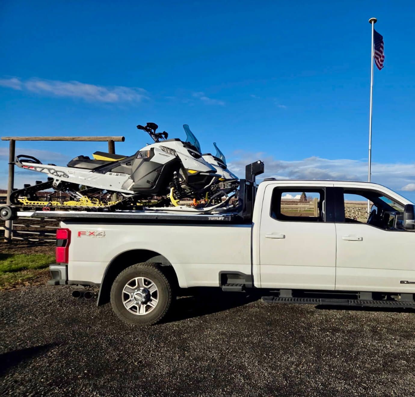 Two snowmobiles loaded on a white truck's TUFFLIFT deck under a blue sky