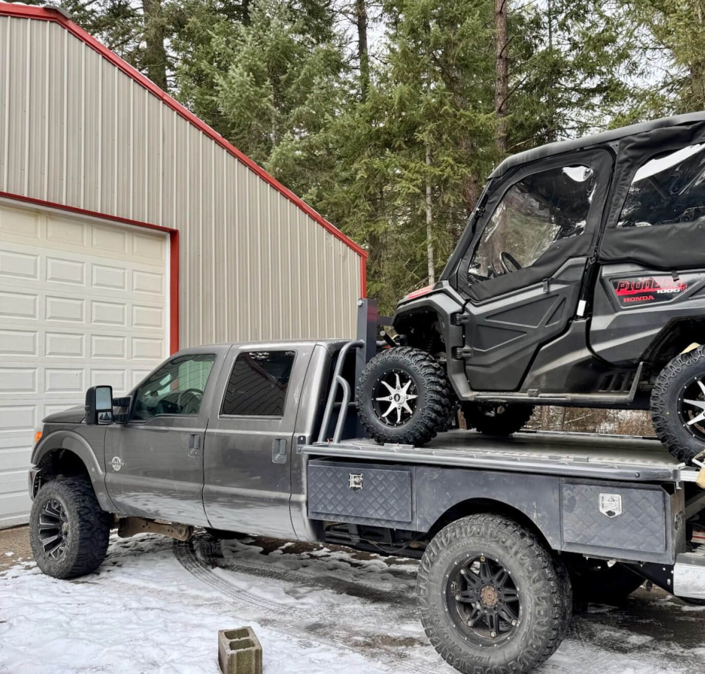 A grey crew-cab truck carrying a black side-by-side on a TUFFLIFT deck outside the shop